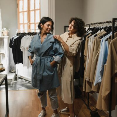 Two women shopping in a boutique, trying on stylish trench coat outfits.